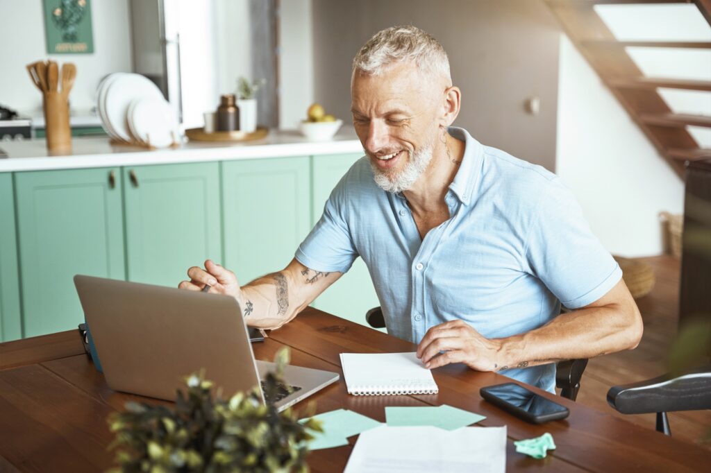 Happy middle aged caucasian man using laptop while sitting at table and working online from